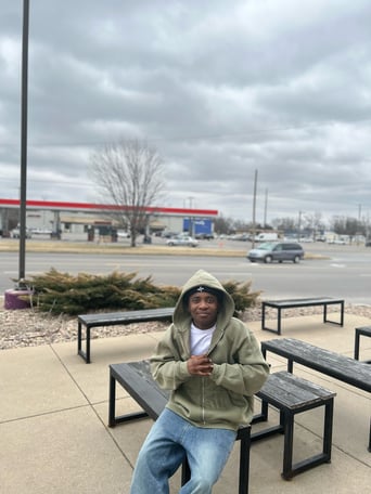 Markcell Tah sits on a picnic bench. He is dressed in a coat and smiling at the camera.